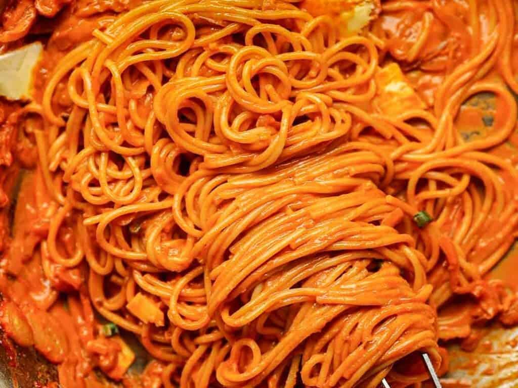 Close-up of spaghetti tangled around a fork, covered in a rich, creamy tomato sauce. The vibrant red-orange sauce coats the strands, with some visible herbs and cheese pieces interspersed. The scene evokes a sense of flavorful indulgence.